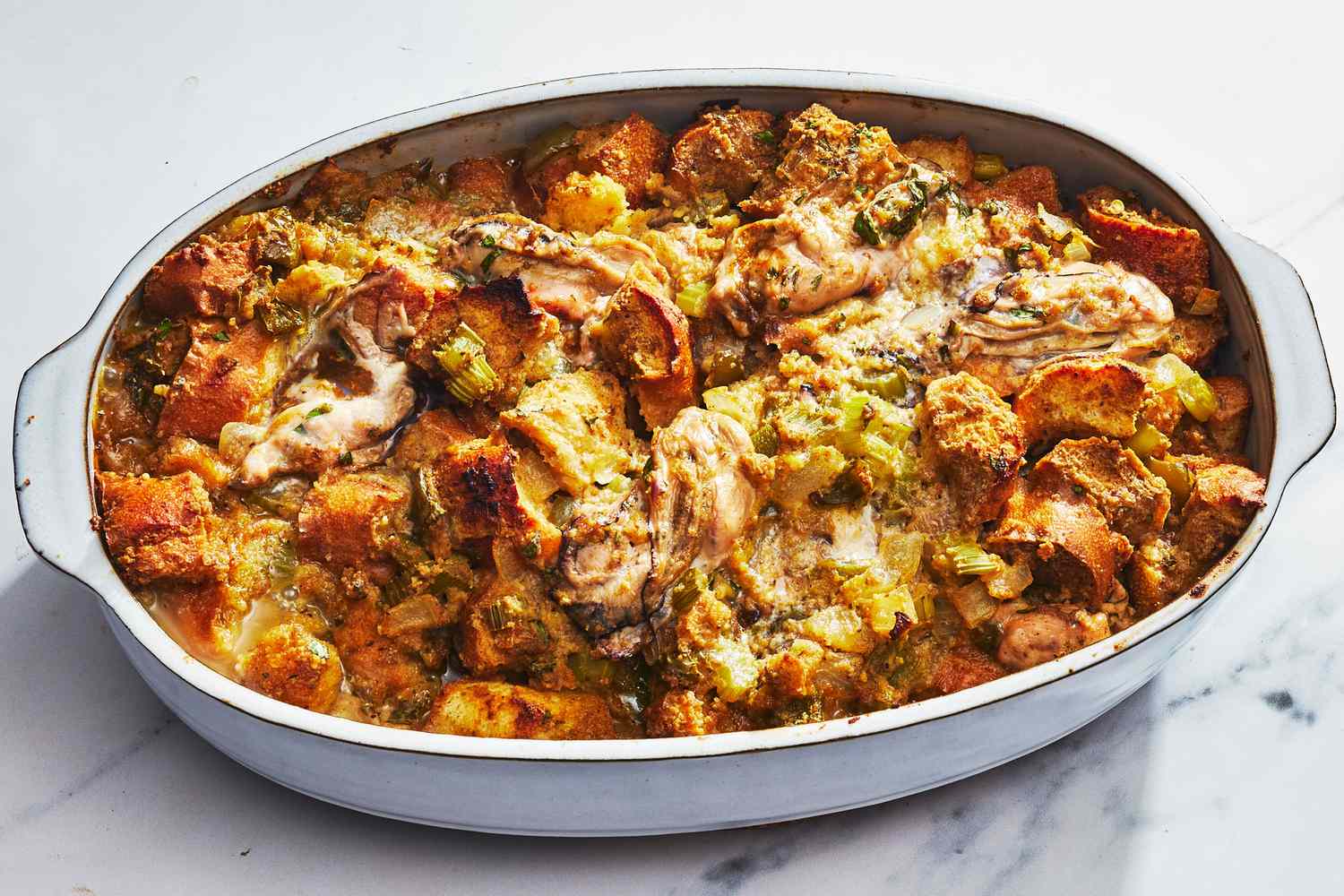 A golden brown baked oyster dressing in a rustic casserole dish on a wooden table with a serving spoon, warm kitchen lighting