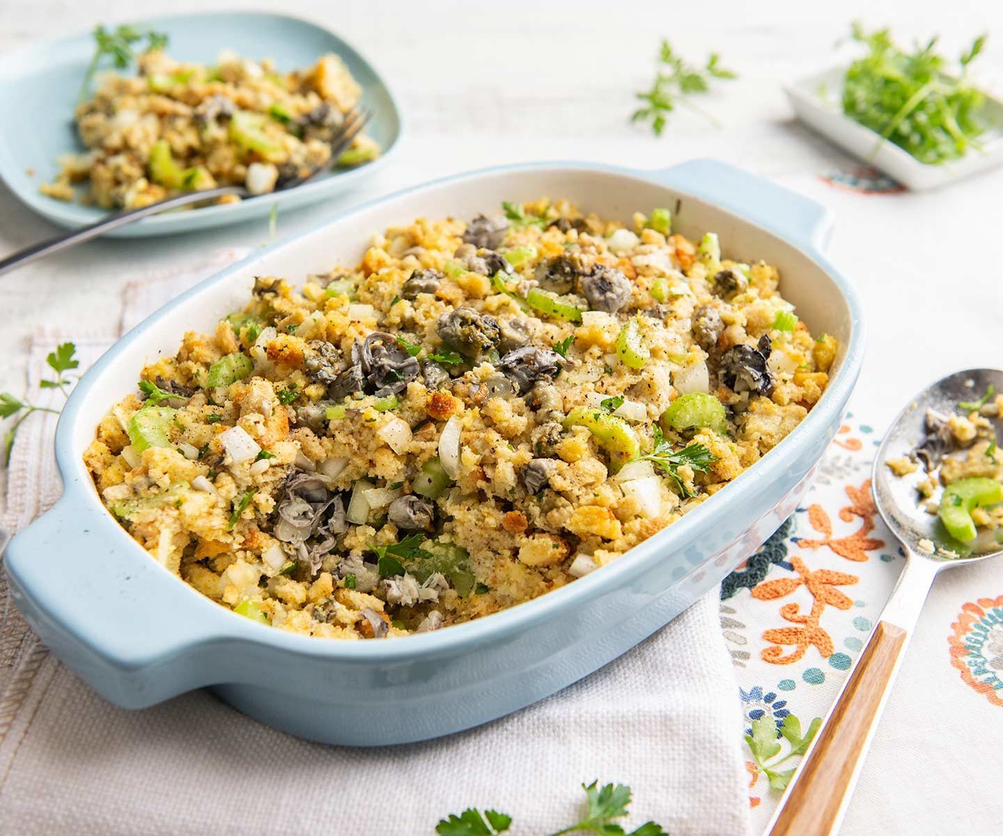 A golden-brown casserole dish of oyster stuffing with crisp edges and visible herbs, sitting on a warm kitchen table with a serving spoon
