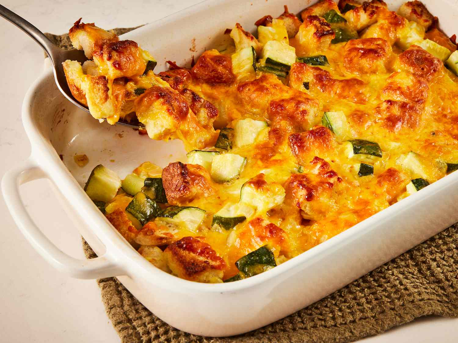 A golden-brown cheesy zucchini casserole in a white ceramic baking dish on a light kitchen counter, with a spoon lifting a creamy portion and a crisp breadcrumb topping