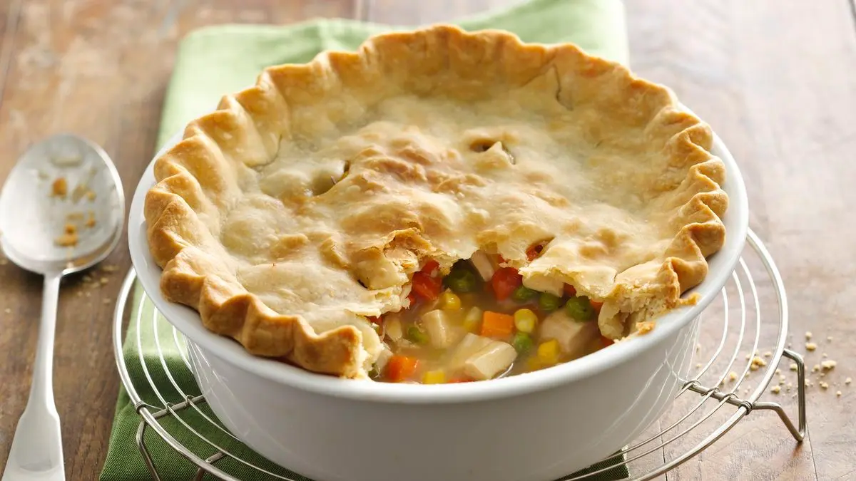 A golden brown chicken pot pie in a ceramic pie dish with a flaky top crust and bubbling filling, on a warm kitchen counter with soft natural light