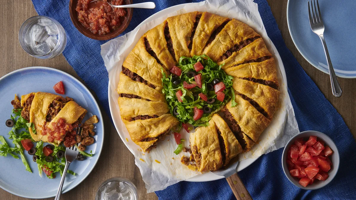 A golden brown crescent roll taco ring on a parchment-lined baking sheet with a small bowl of salsa in the center, warm kitchen lighting