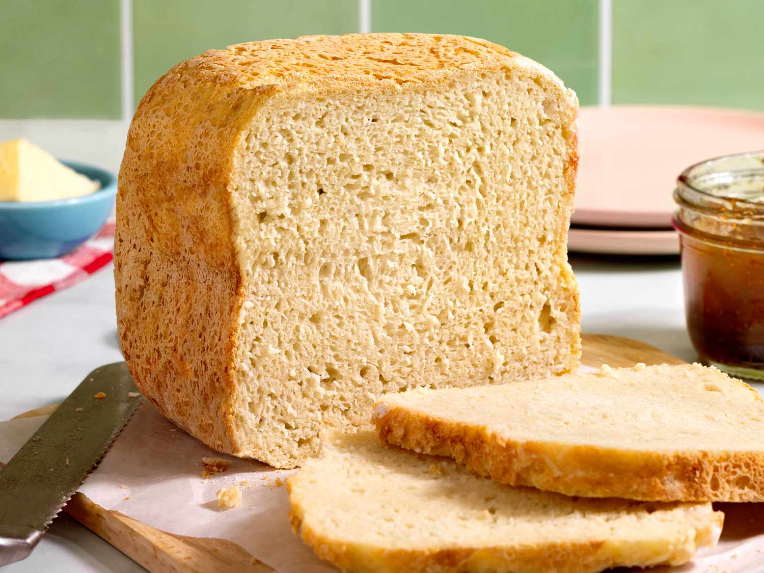A golden brown gluten free sandwich bread loaf resting on a wooden cutting board with a few slices cut, crumbs visible, on a bright kitchen counter