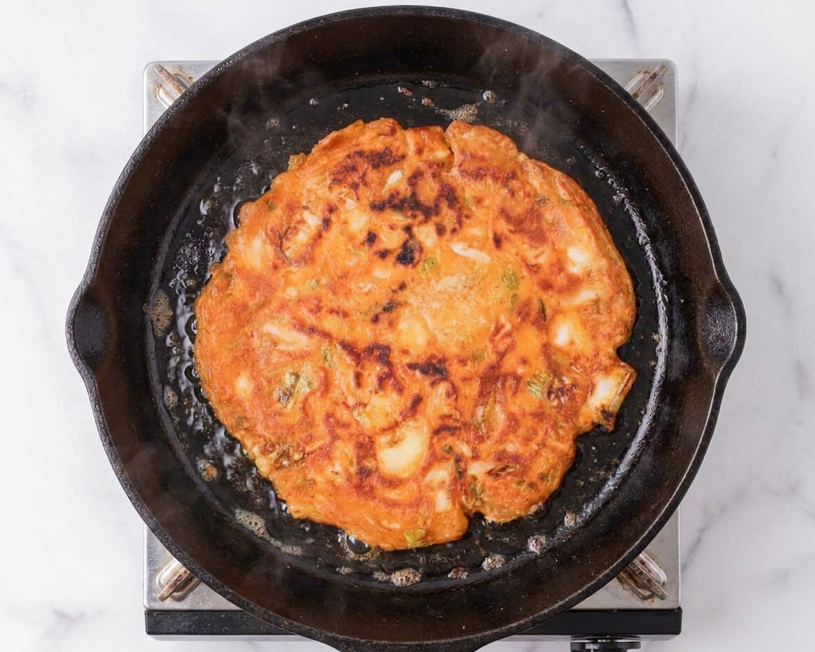 A golden brown kimchi pancake cooking in a cast iron skillet on a home stove, with crisp lacy edges and visible kimchi pieces