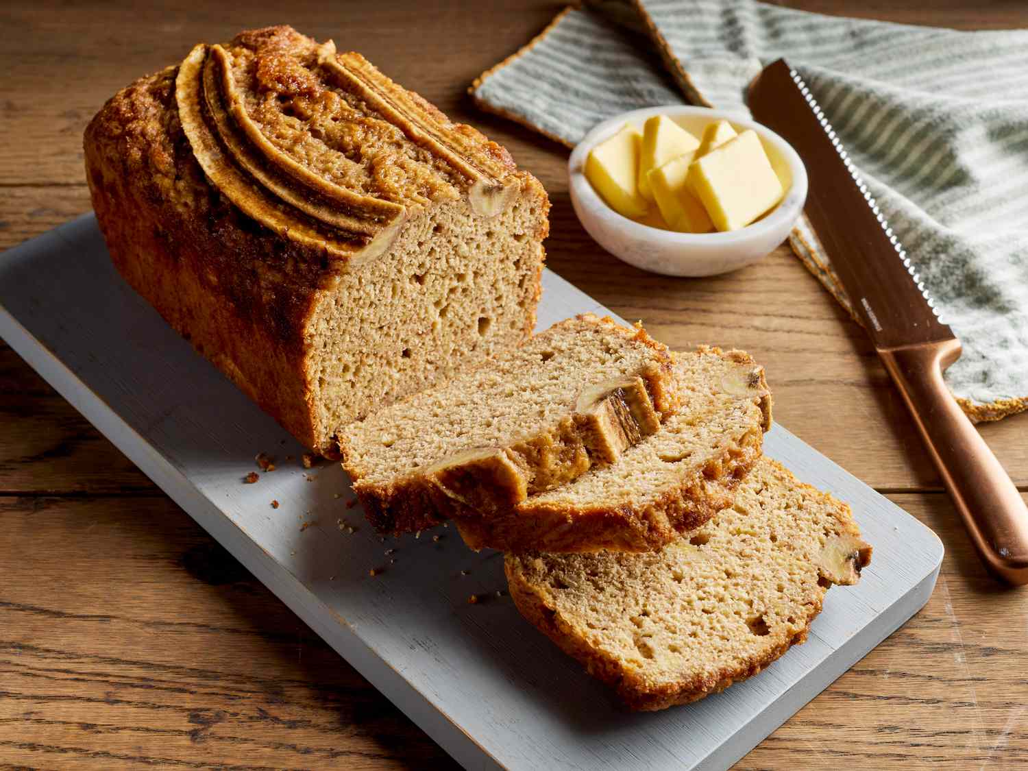 A golden brown loaf of banana bread cooling on a wire rack on a light kitchen counter, with a few slices cut to show a moist crumb