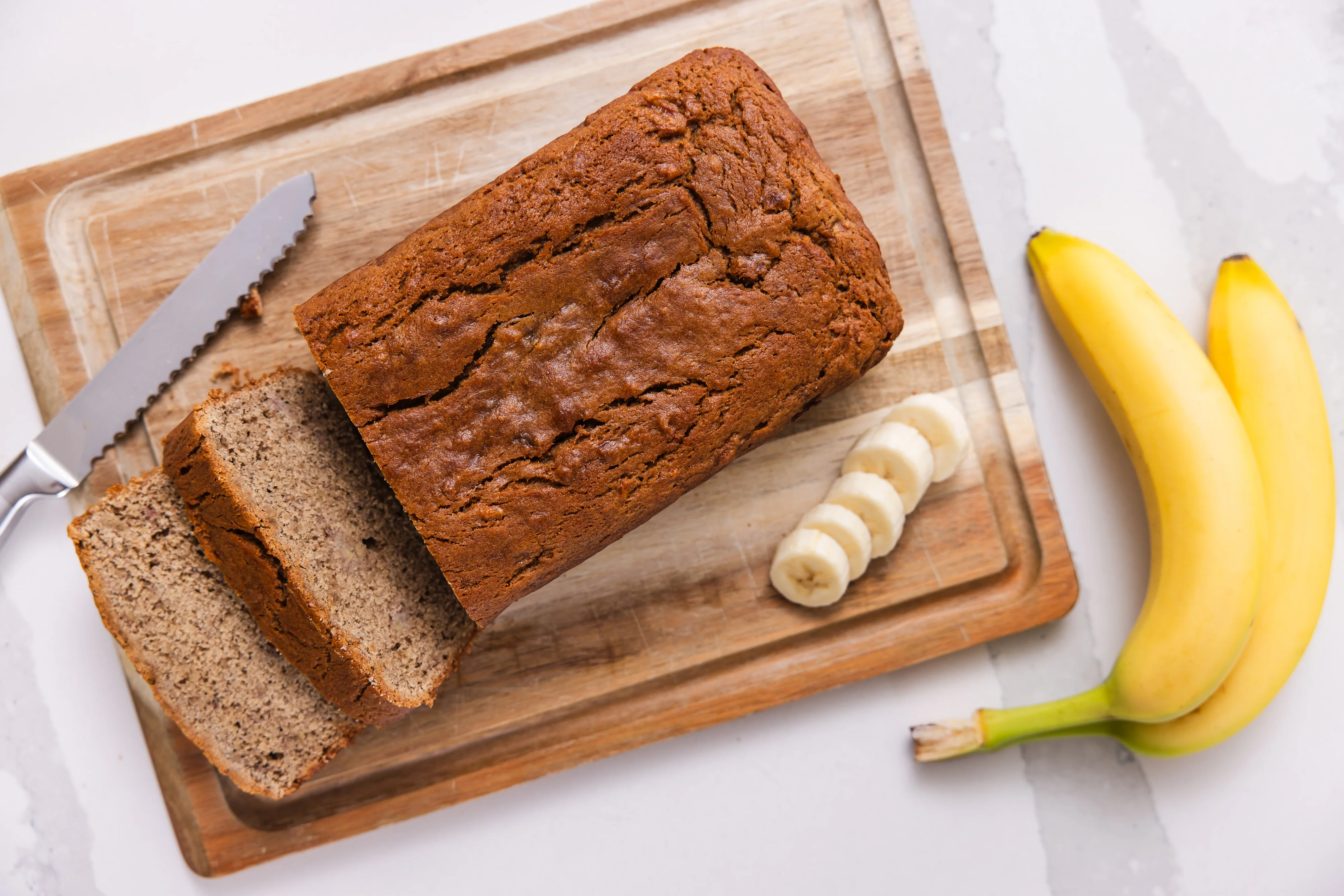 A golden brown loaf of gluten-free banana bread sliced on a wooden cutting board with a few banana slices nearby on a sunlit kitchen counter