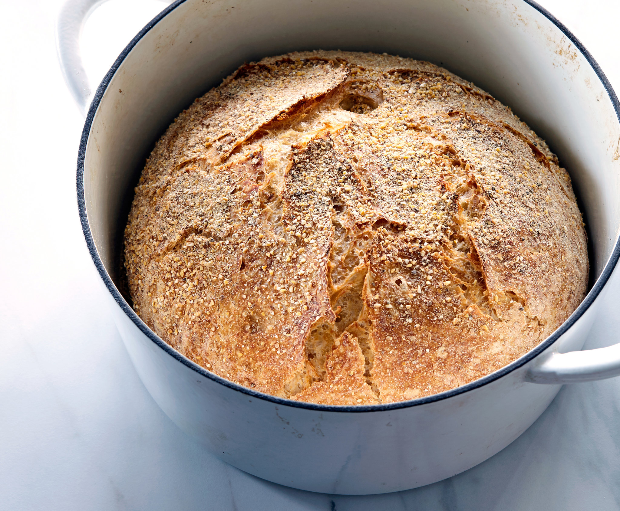 A golden brown no-knead round loaf with a crackly crust sitting on parchment paper beside an open cast iron Dutch oven on a clean kitchen counter in soft natural light