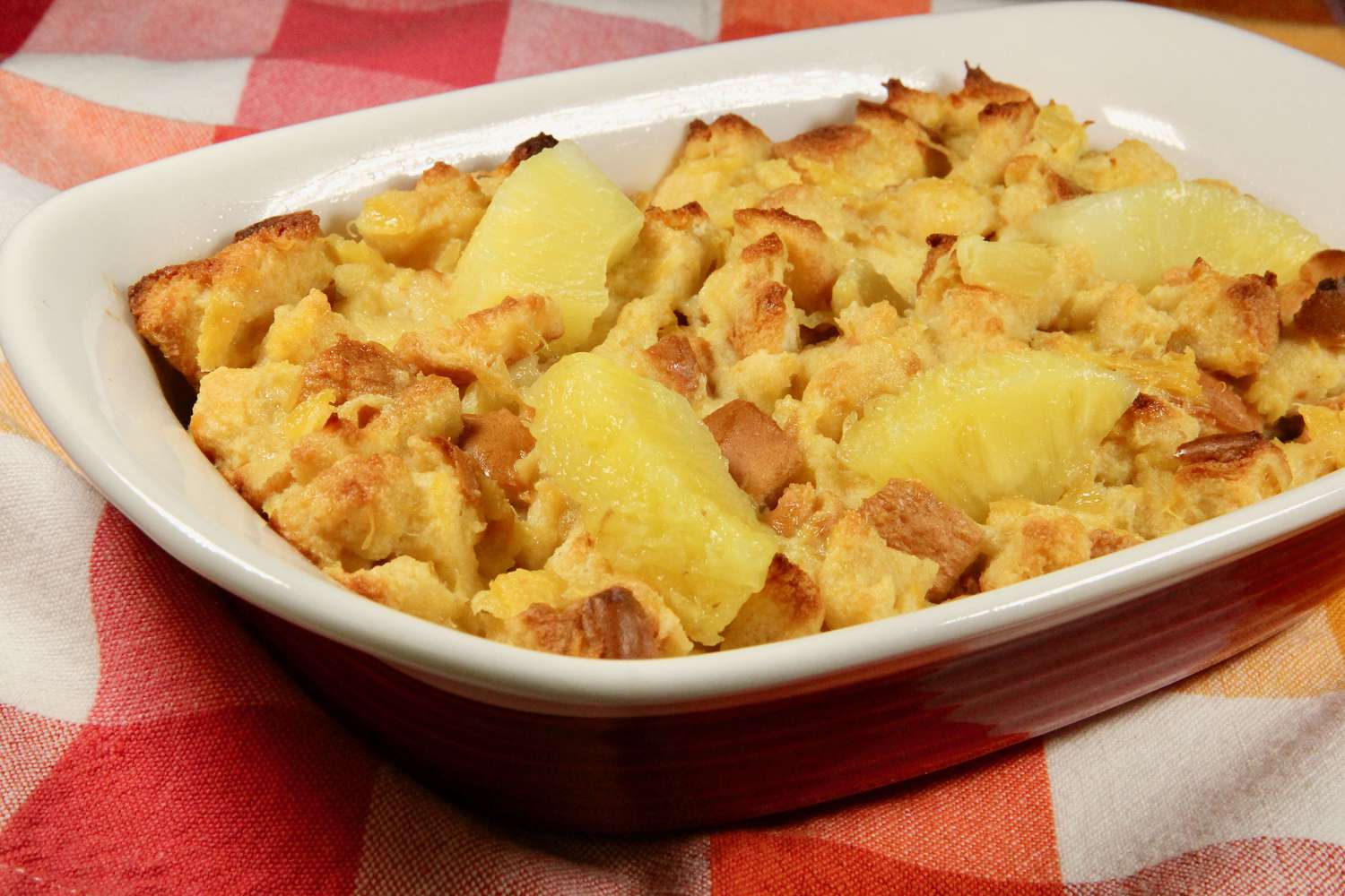 A golden-brown pineapple stuffing baked in a glass casserole dish on a wooden table, with lightly crisp edges and a soft, custardy center