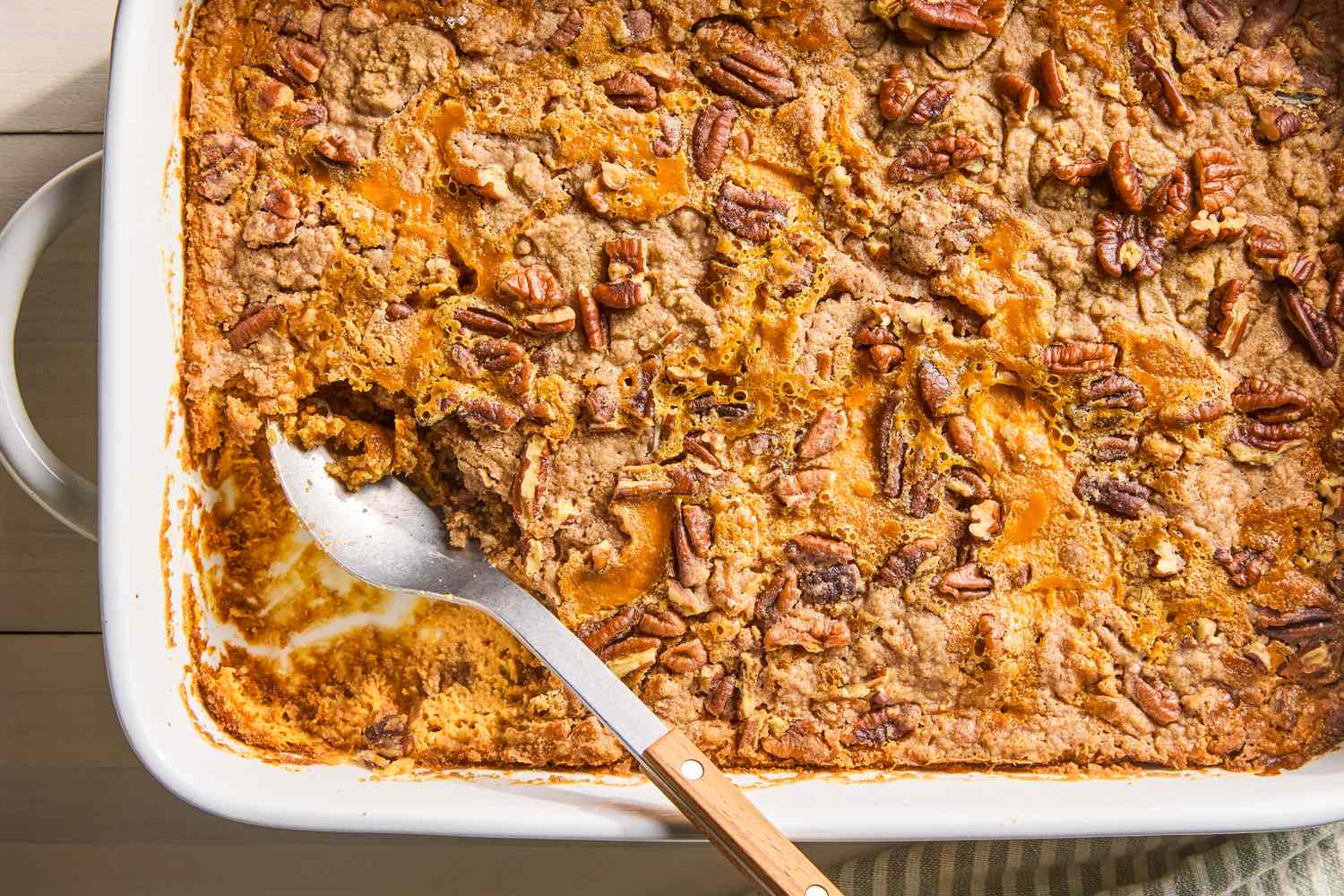 A golden brown pumpkin dump cake in a glass baking dish with a crisp buttery topping and a serving spoon resting in the corner, on a warm-toned kitchen counter