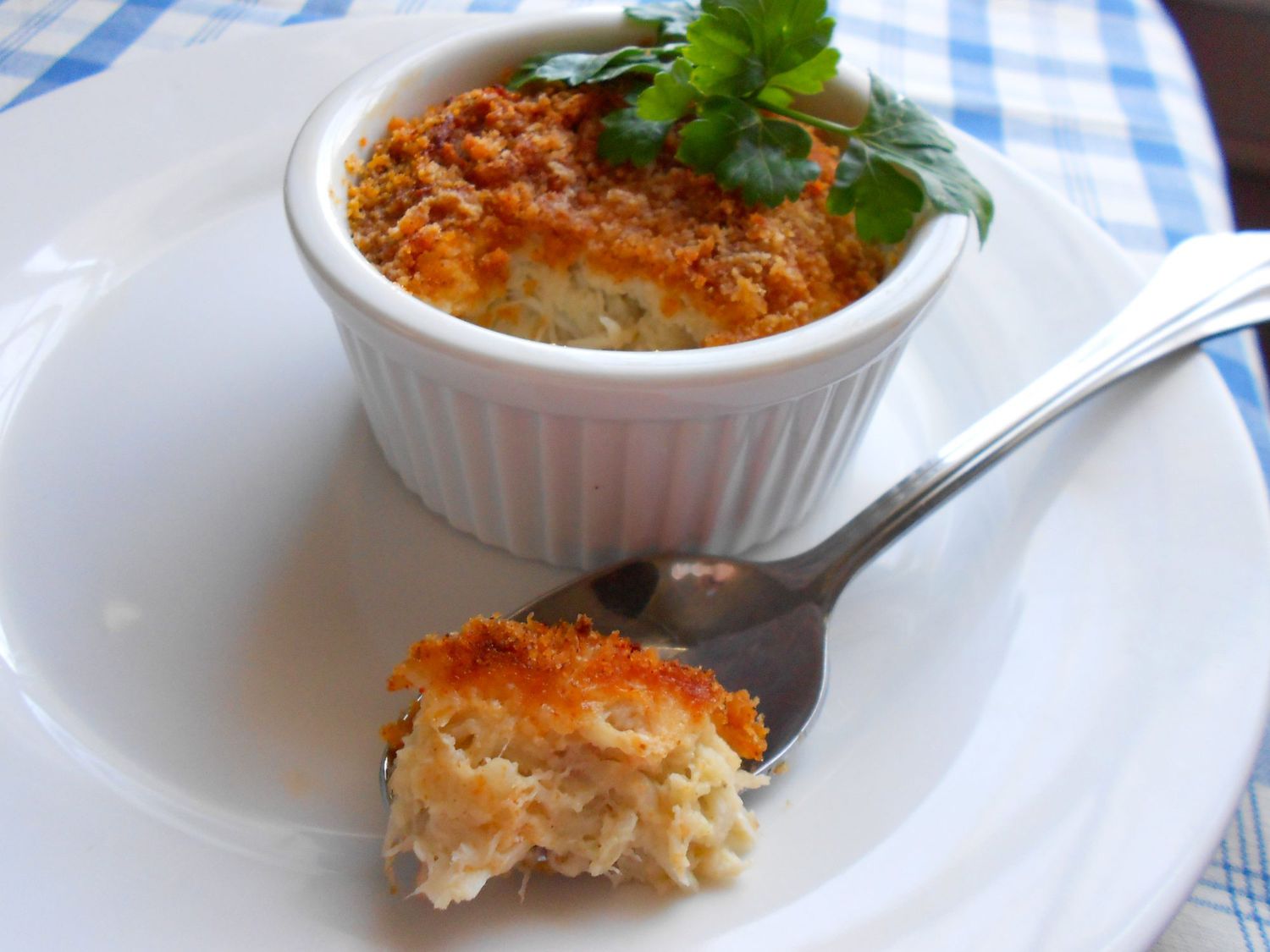 A golden-brown ramekin of crab imperial with a crisp breadcrumb topping on a neutral dinner plate, with a lemon wedge and parsley nearby on a stone countertop in warm kitchen light