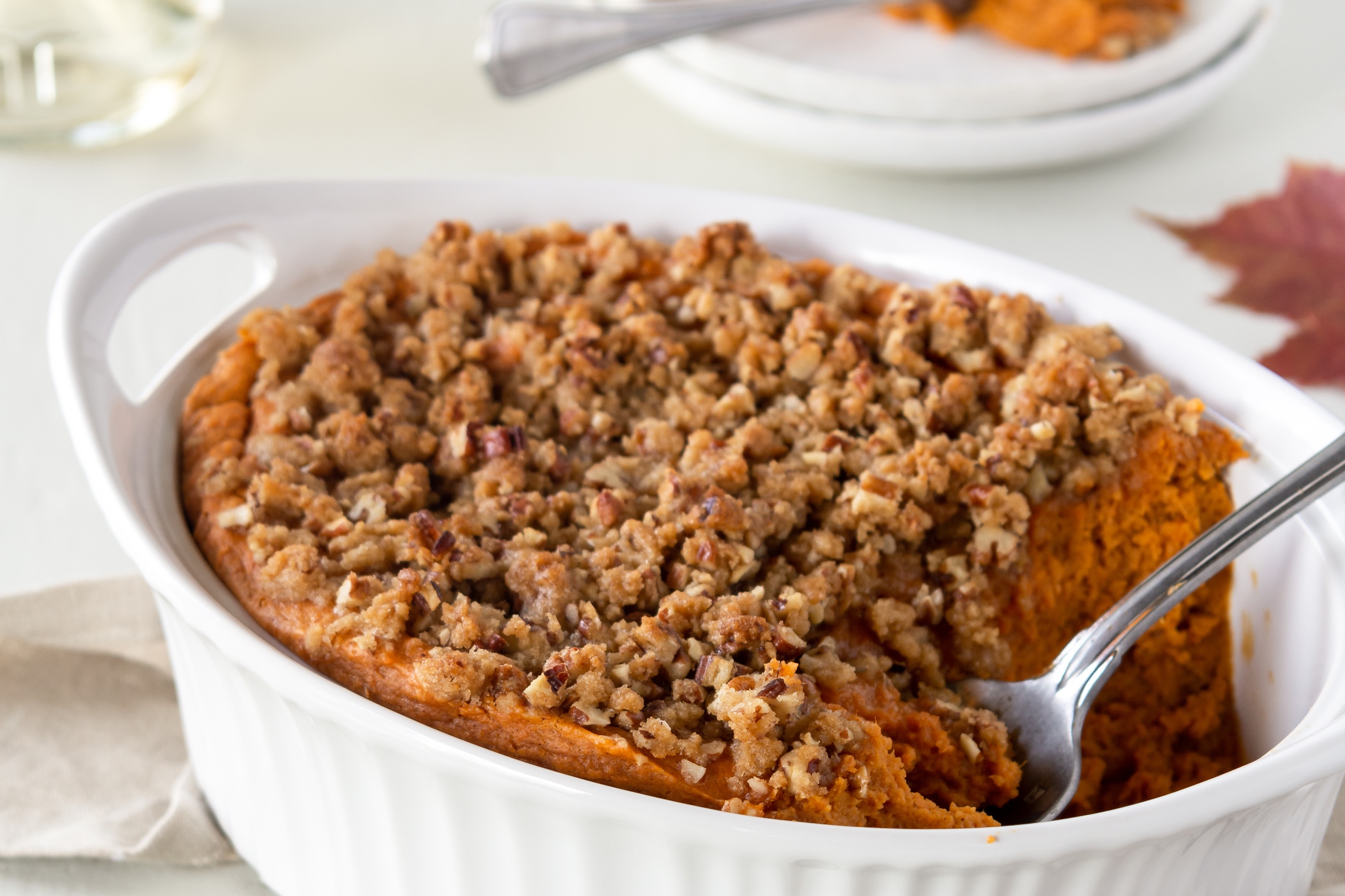 A golden-brown sweet potato soufflé in a white ceramic baking dish with a crisp pecan streusel topping, set on a warm-toned kitchen counter with soft natural light
