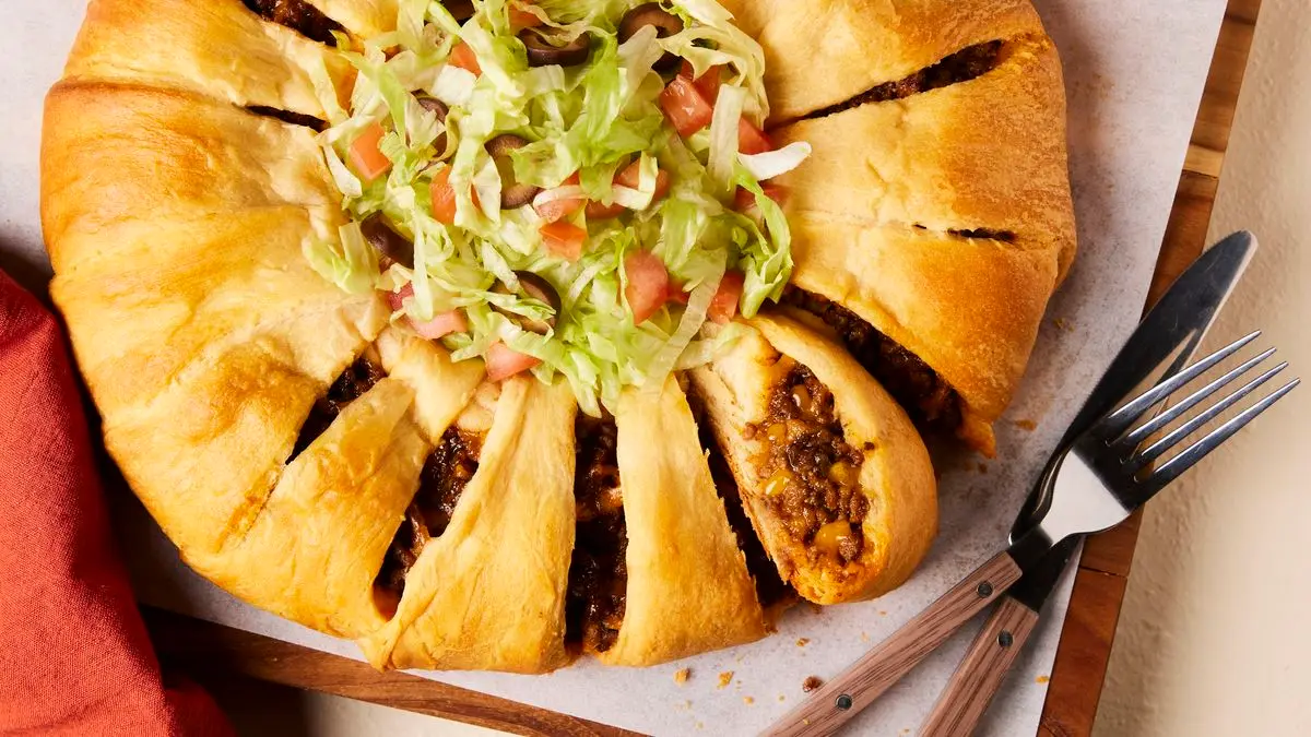 A golden brown taco ring made with crescent roll dough on a parchment-lined baking sheet, sliced into wedges with small bowls of salsa and sour cream nearby on a cozy kitchen table