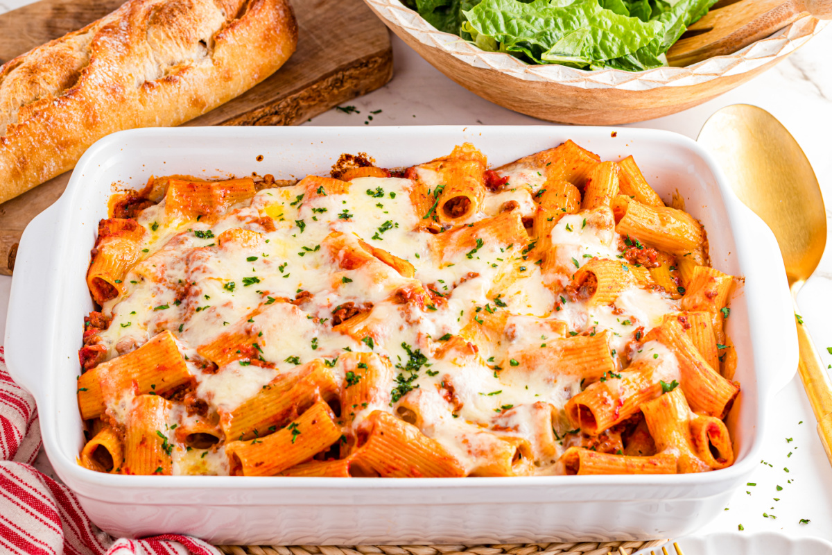 A golden, bubbly baked rigatoni in a white ceramic baking dish on a warm kitchen table, with browned cheese on top and a spoon scooping out a saucy portion