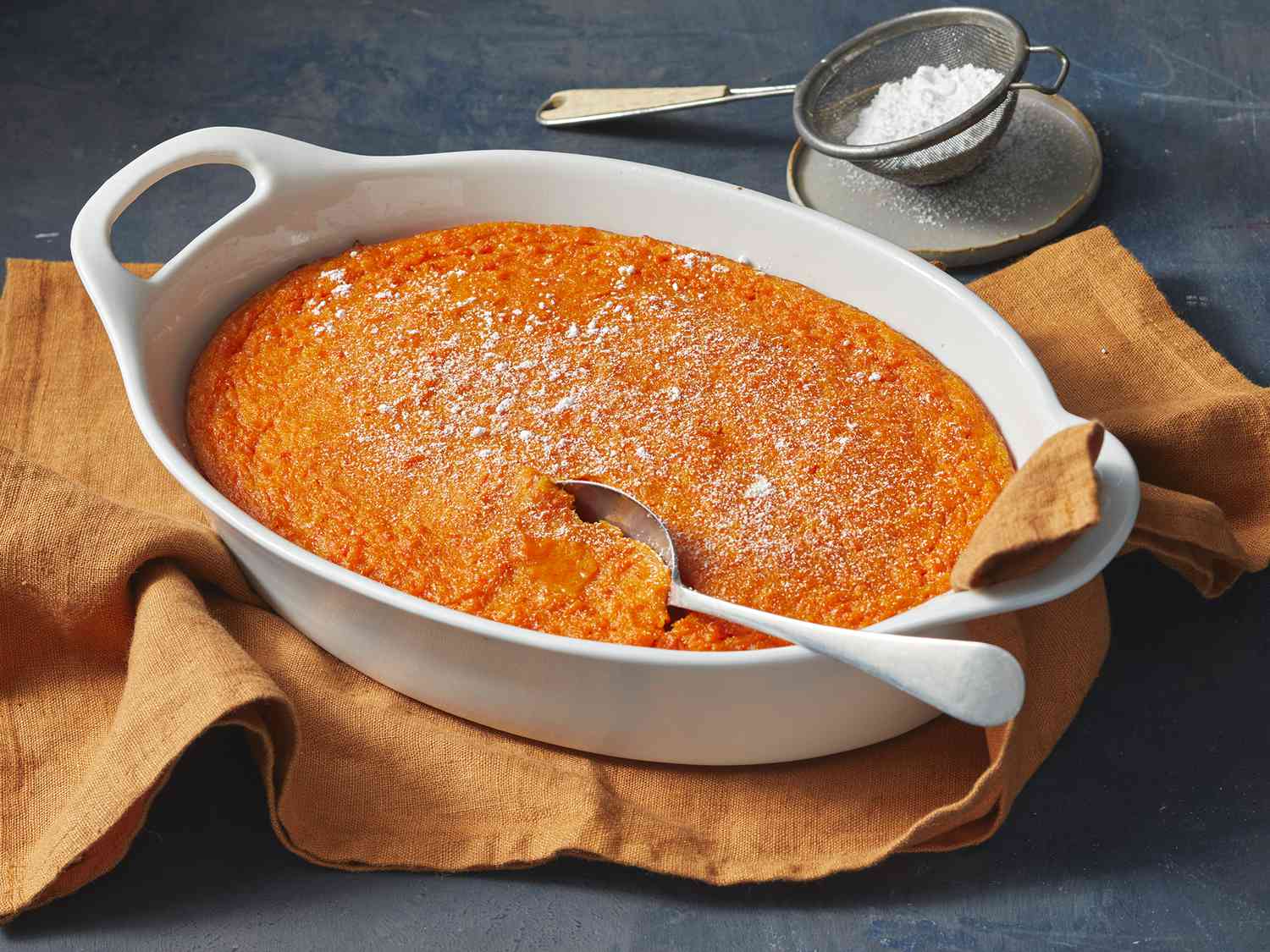 A golden carrot soufflé in a white ceramic baking dish on a light stone countertop, with browned edges and a soft, slightly puffed center