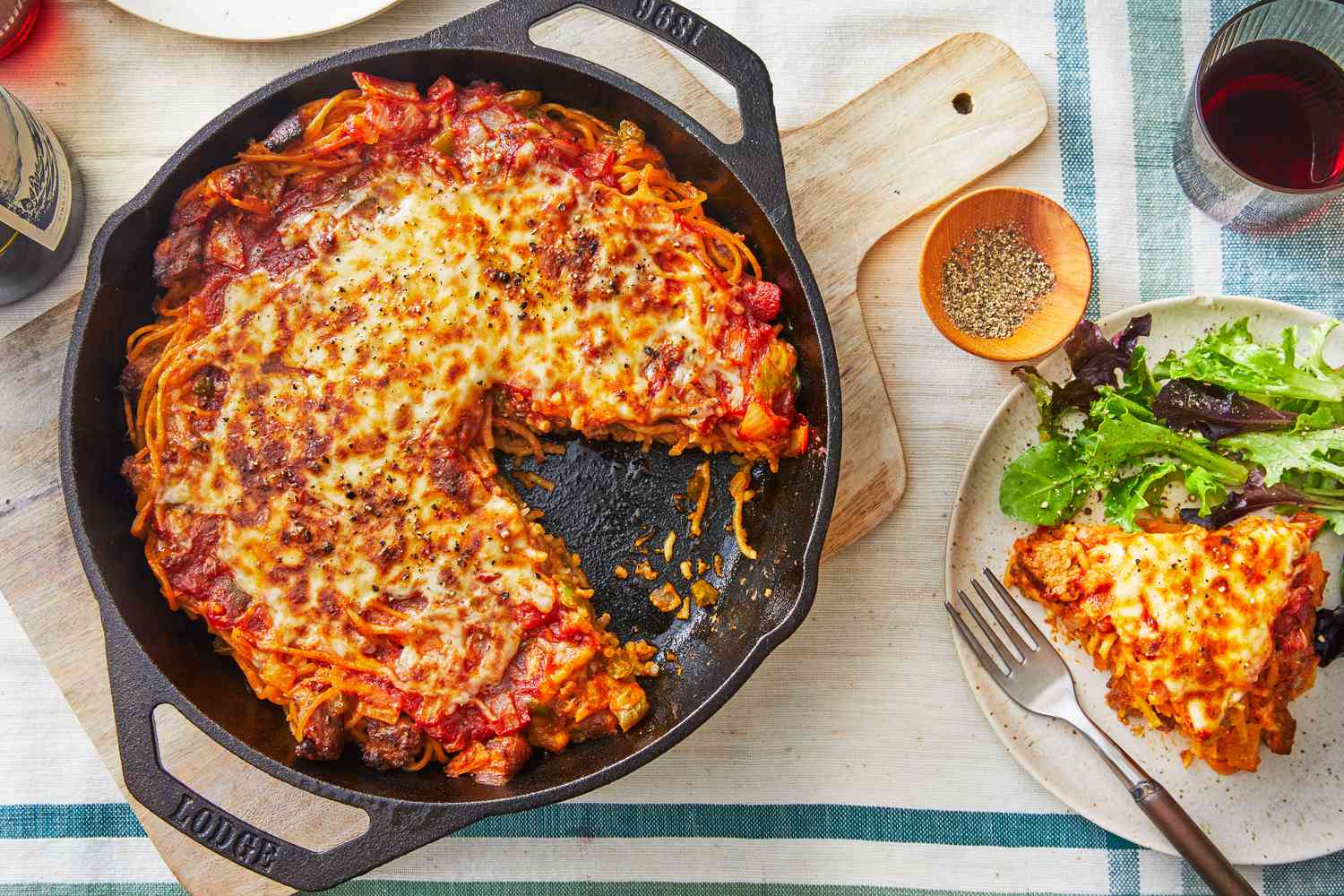 A golden, cheesy spaghetti pie in a cast iron skillet on a wooden table, with one slice lifted to show the layered spaghetti crust and meat sauce