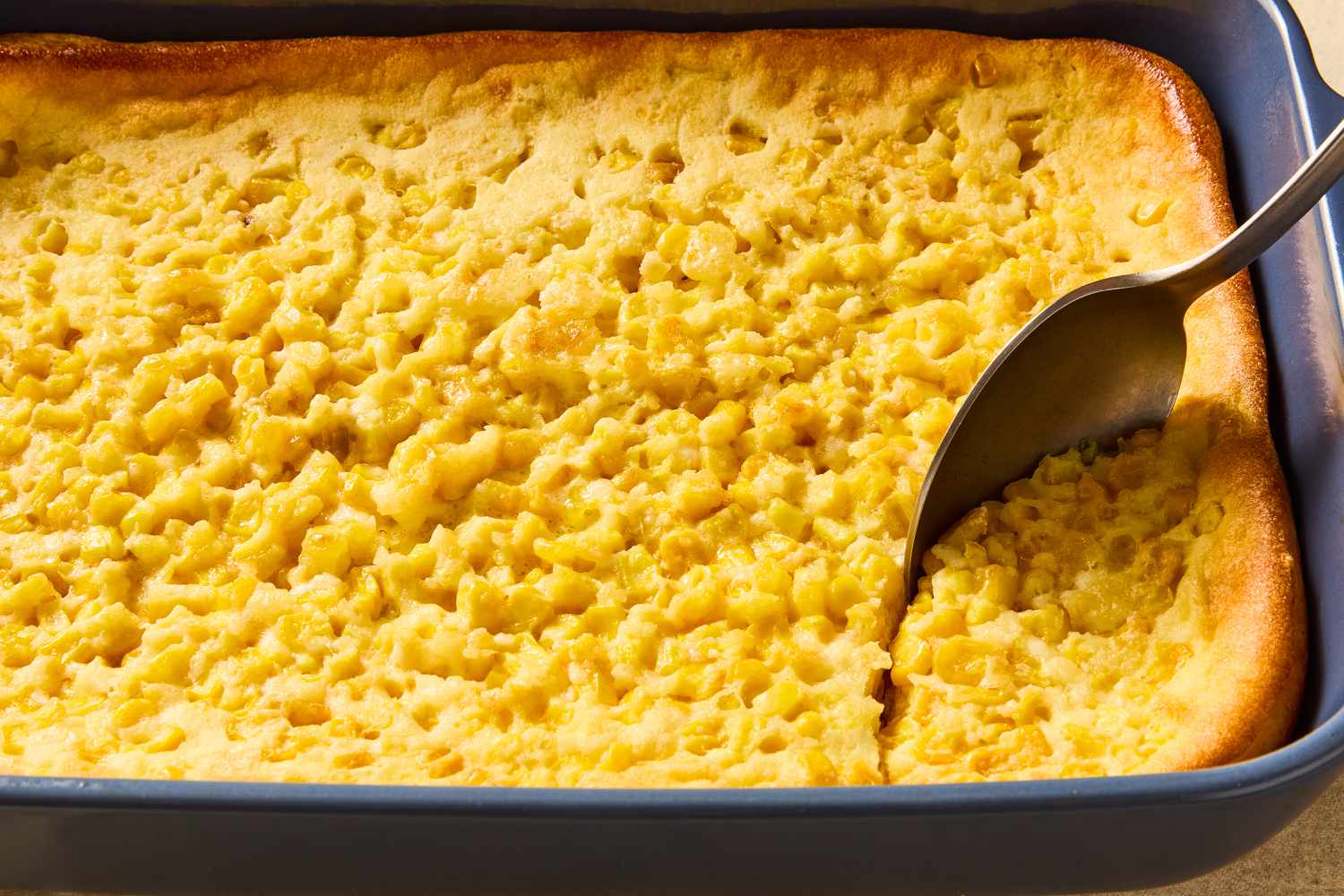 A golden corn souffle casserole in a white baking dish on a warm kitchen table, with a serving spoon and soft natural window light
