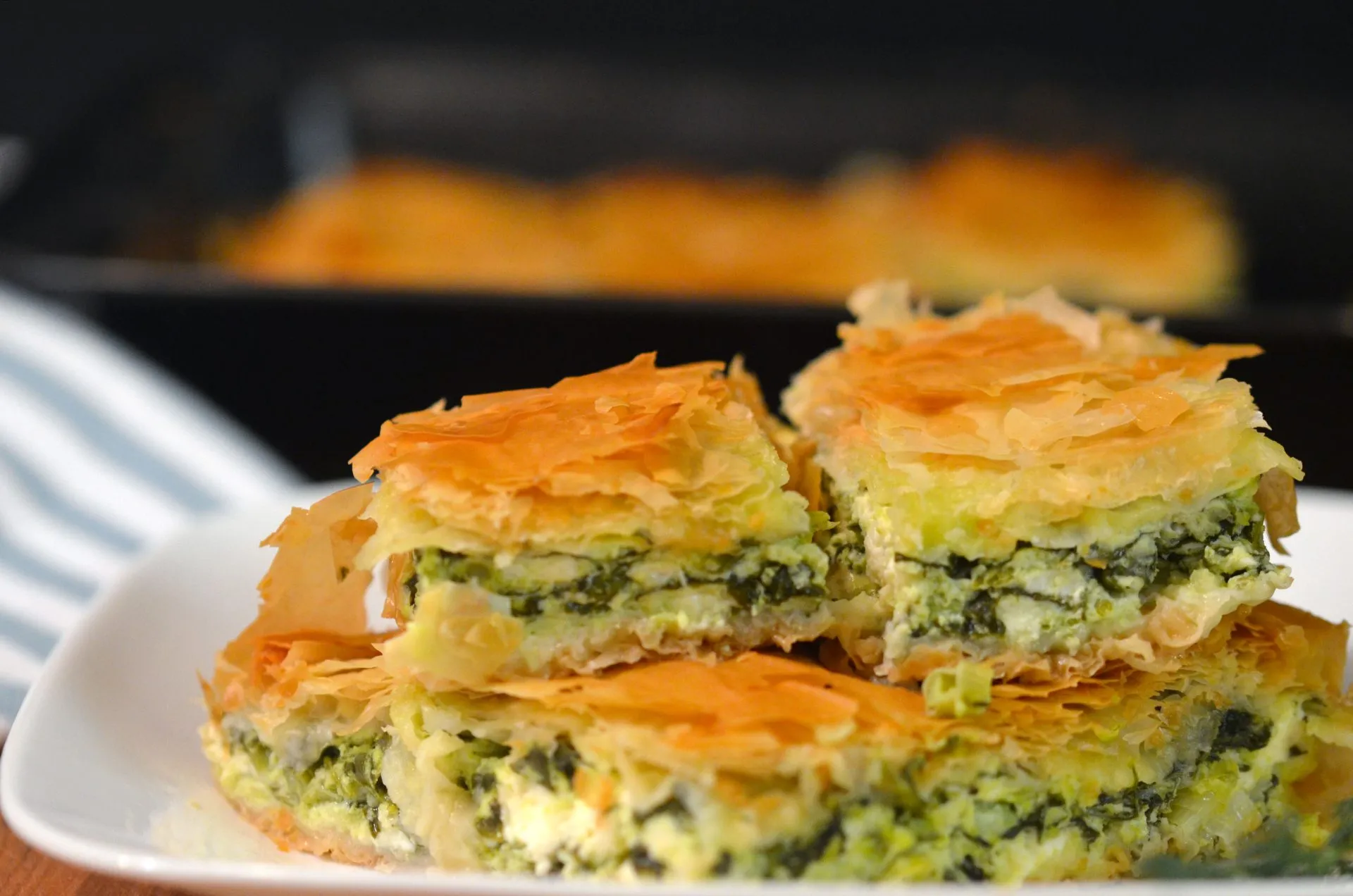 A golden, flaky spinach pie slice on a white plate with visible layers of phyllo and a creamy spinach and feta filling, set on a warm-toned kitchen table
