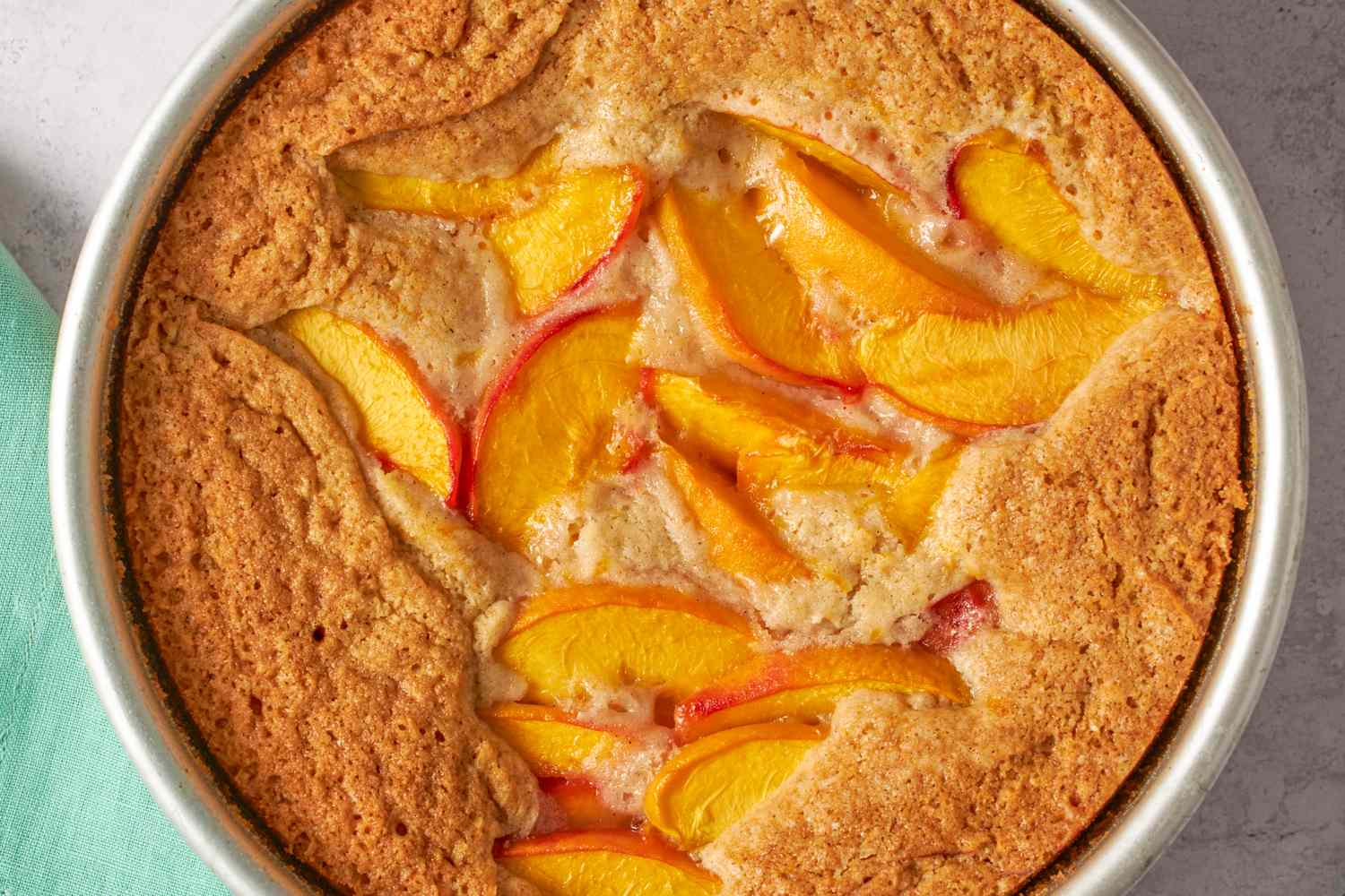 A golden fresh peach cake baked in a round pan on a light kitchen counter, topped with cinnamon sugar and scattered peach slices, natural window light