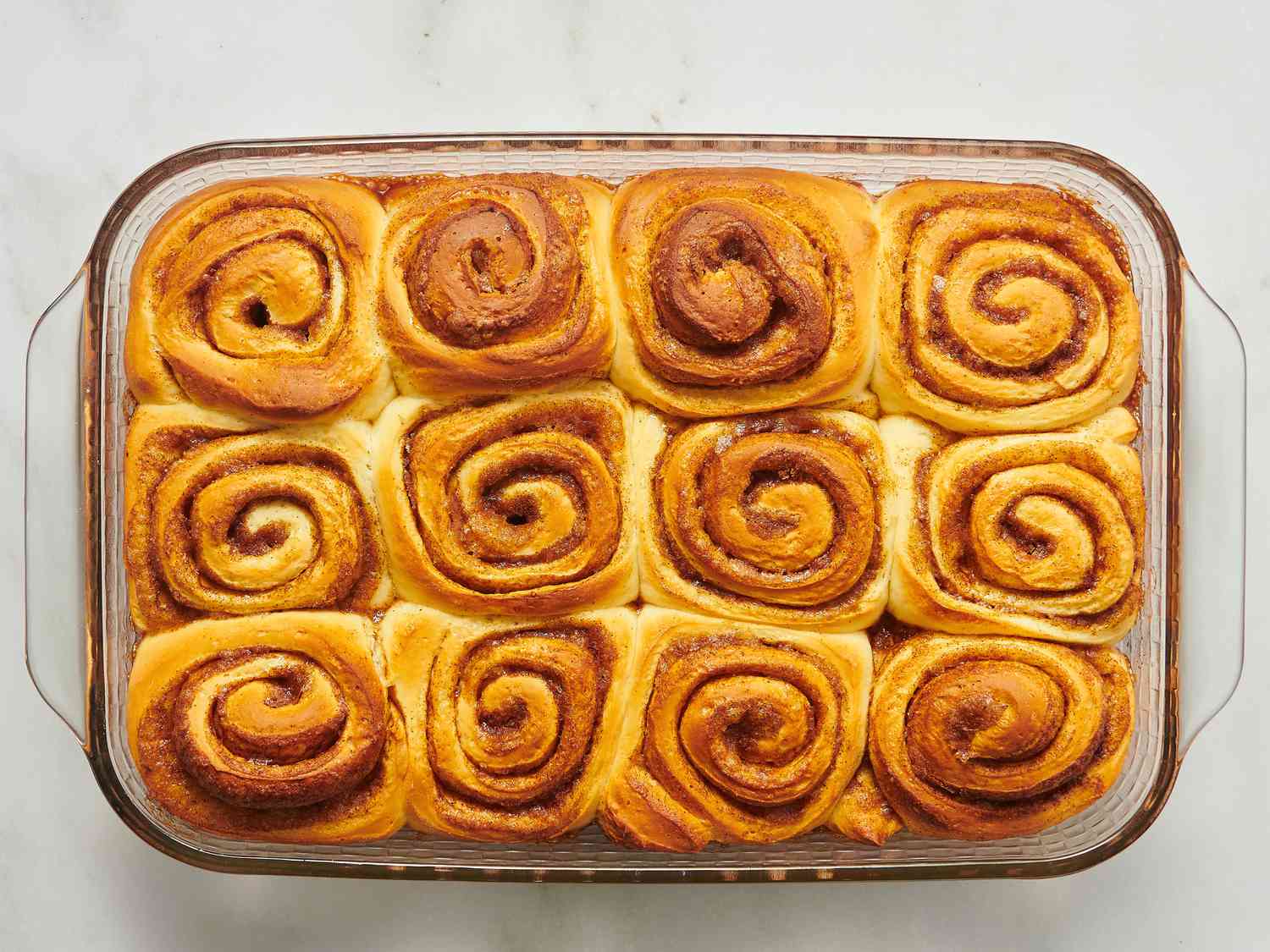 A golden pan of freshly baked cinnamon buns in a glass baking dish on a light kitchen counter, icing starting to melt into the warm spirals