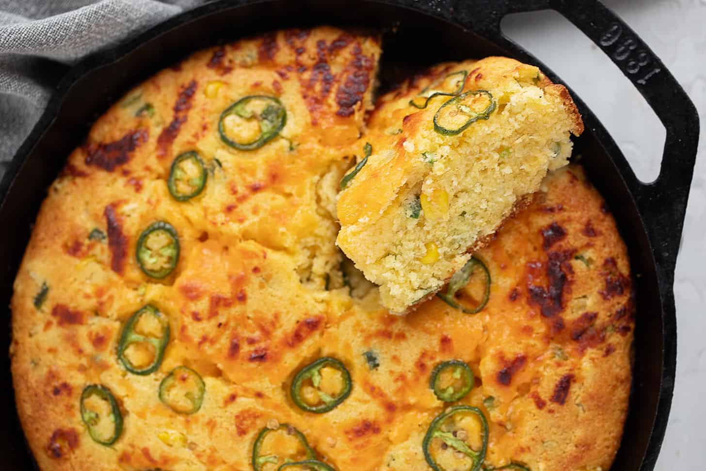 A golden skillet of jalapeño cheddar cornbread with crisp edges and sliced jalapeños on top, sitting on a rustic kitchen counter in warm natural light