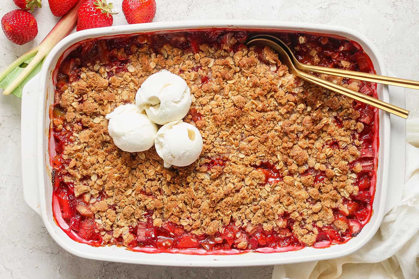A golden strawberry rhubarb crisp baked in a 9x13 glass pan on a light kitchen counter, with bubbling red fruit visible around the edges