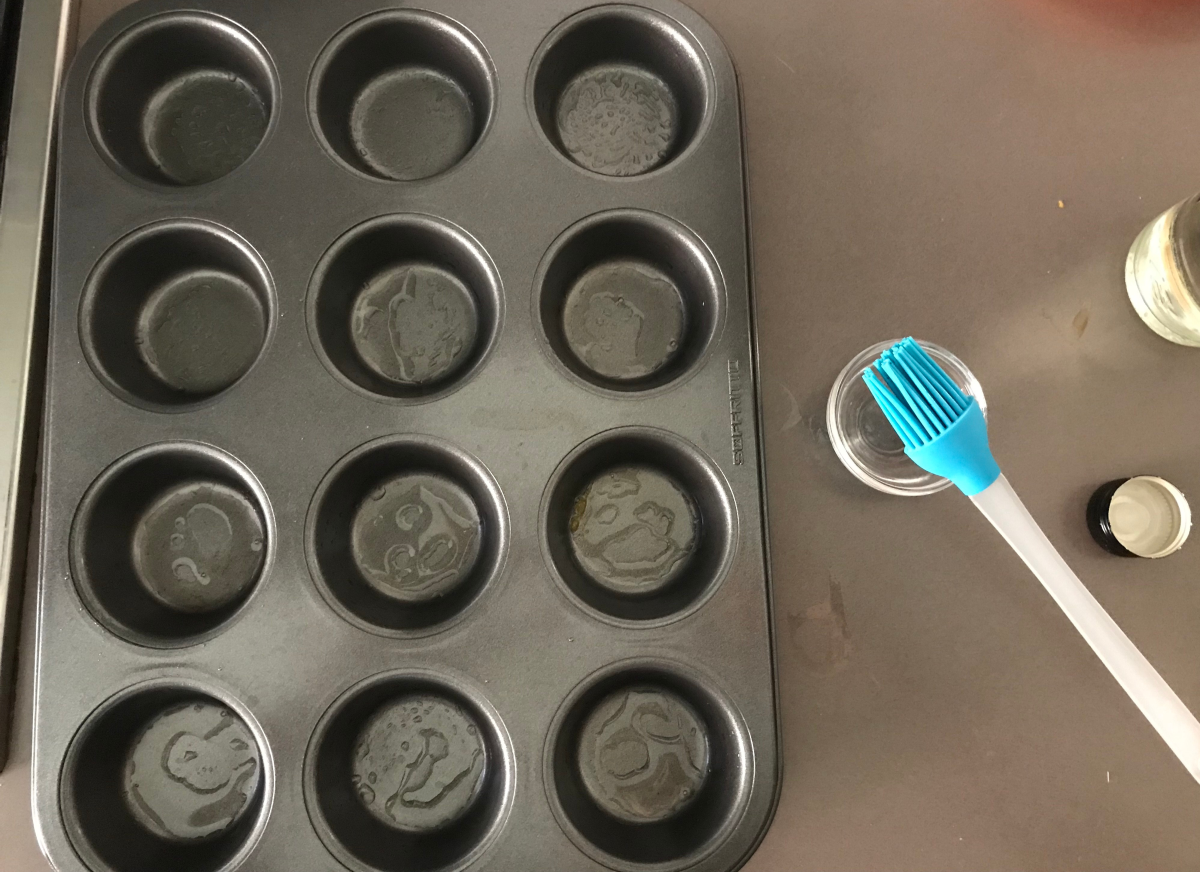 A greased metal muffin tin set on a sheet pan on a home kitchen counter under warm light