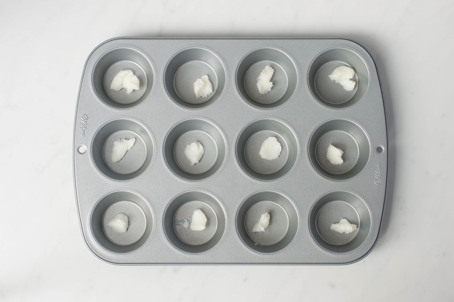 A greased nonstick 12-cup muffin tin on a light stone countertop with a small pastry brush and a bottle of oil nearby