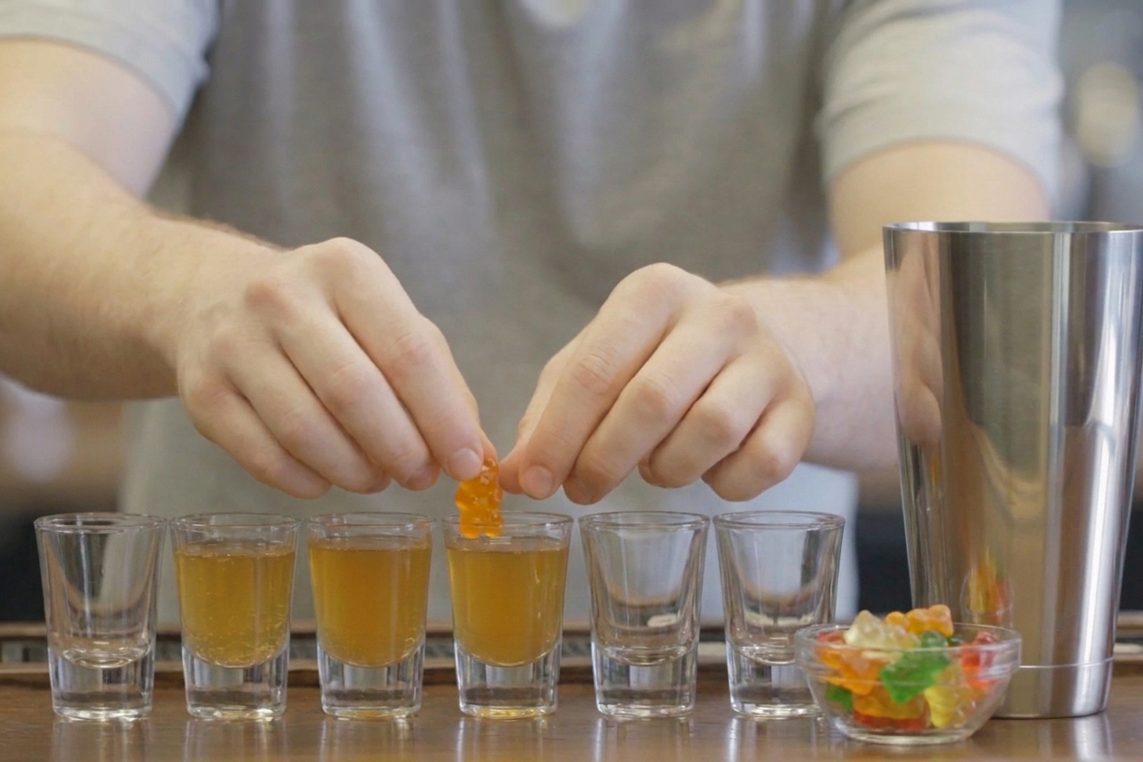 A gummy bear garnish being placed onto a filled shot glass of golden-orange shooter on a kitchen counter