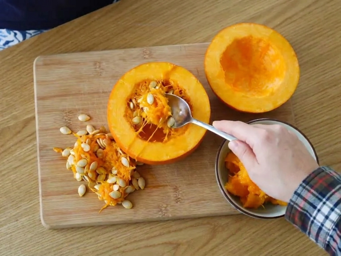 A halved sugar pumpkin on a cutting board while the seeds and stringy pulp are being scooped out with a spoon