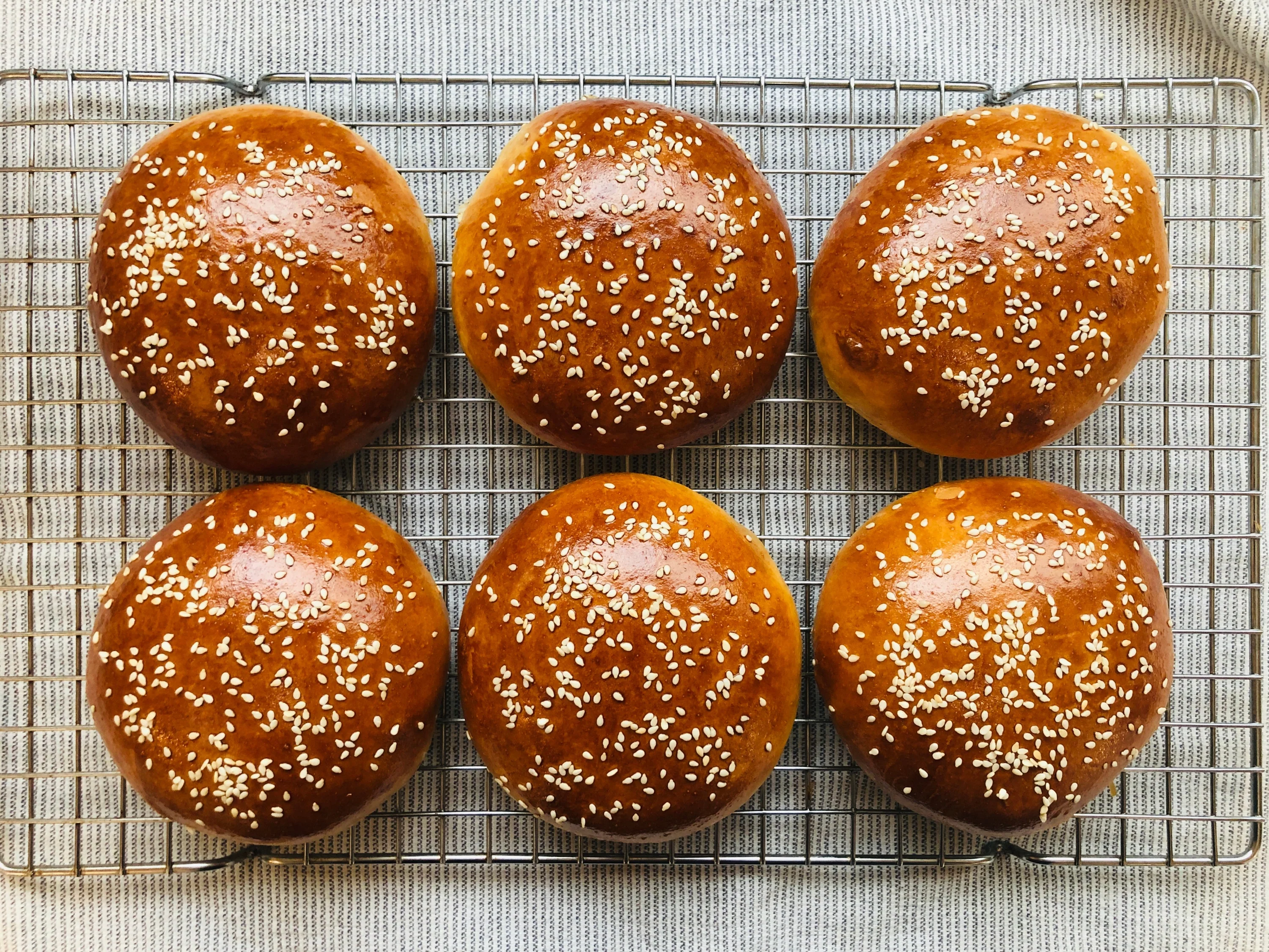 A hand brushing egg wash over unbaked hamburger roll dough rounds on a parchment-lined baking sheet in a home kitchen