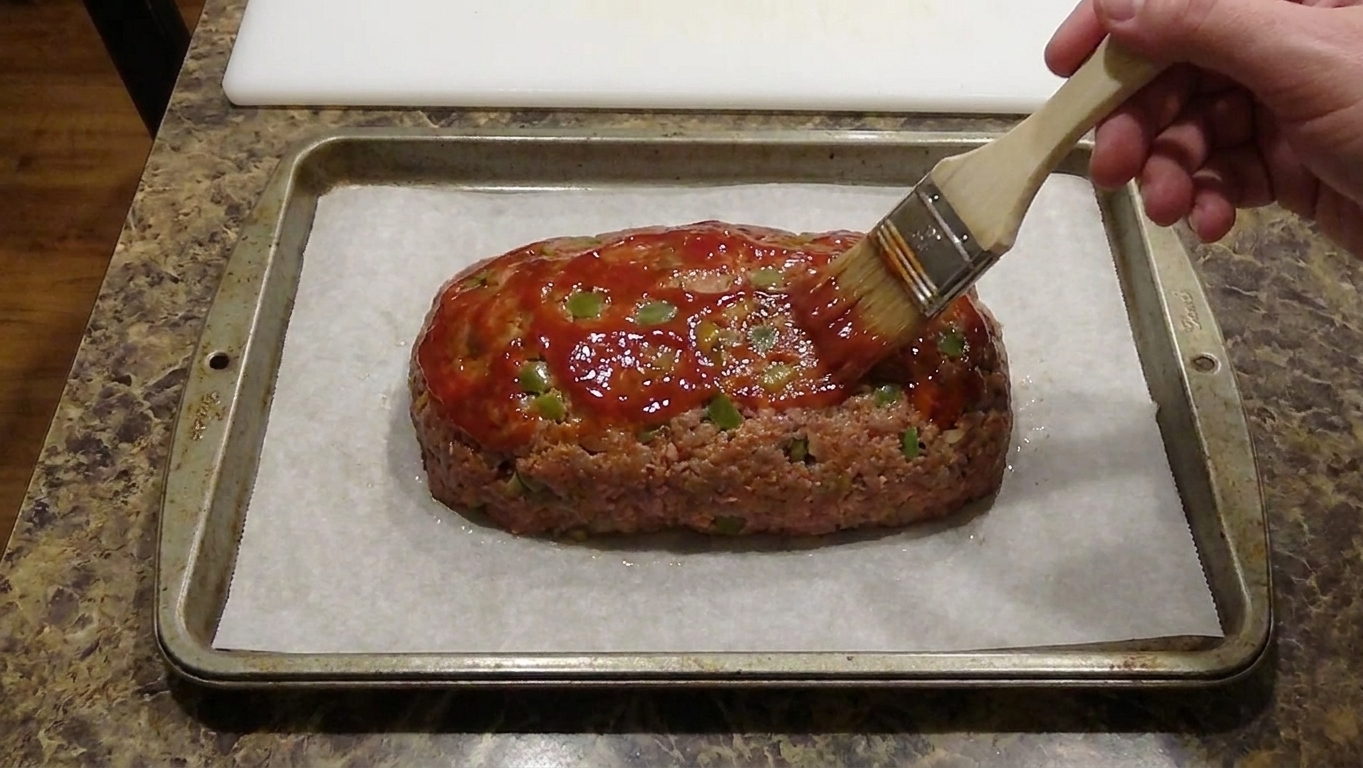 A hand brushing ketchup glaze over a partially baked meatloaf on a baking sheet
