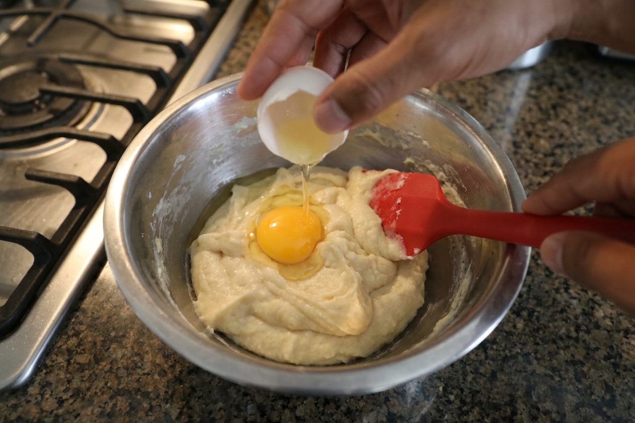 A hand cracking an egg into a bowl of choux dough while mixing, with a rubber spatula and clean countertop