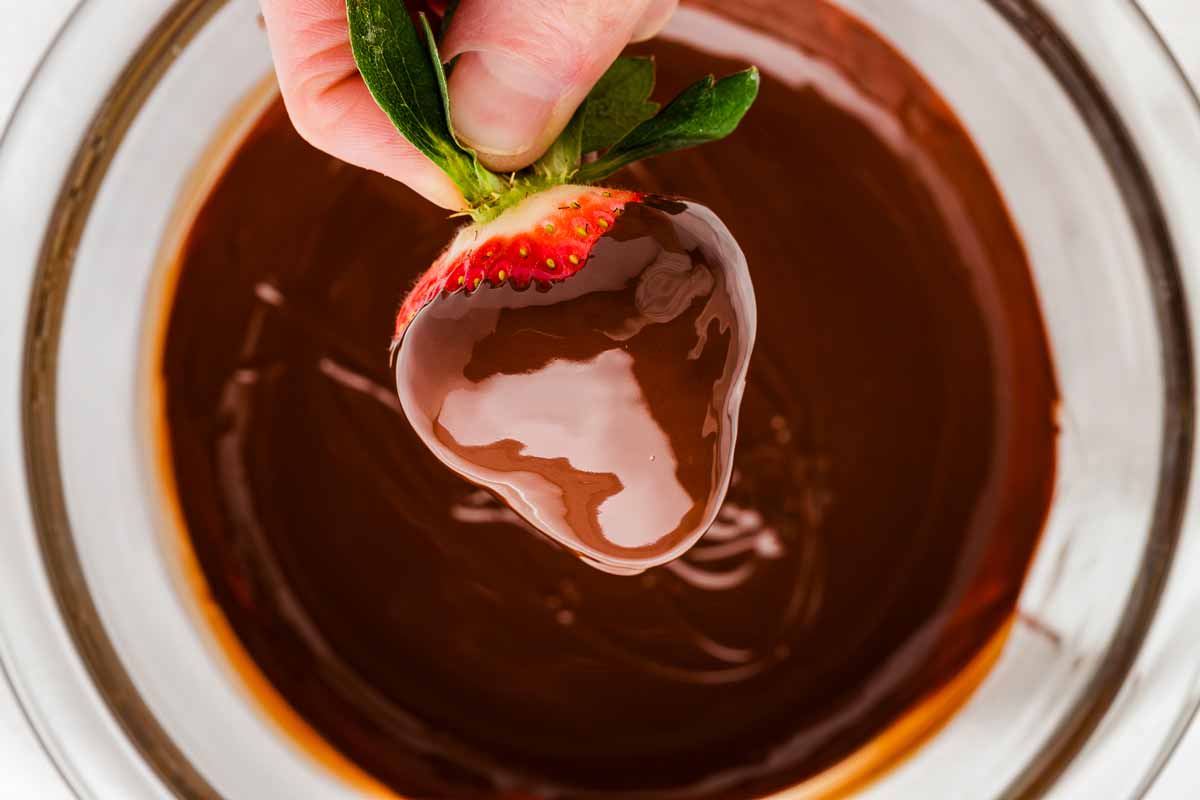 A hand dipping a strawberry into a bowl of smooth melted chocolate with chocolate dripping back into the bowl