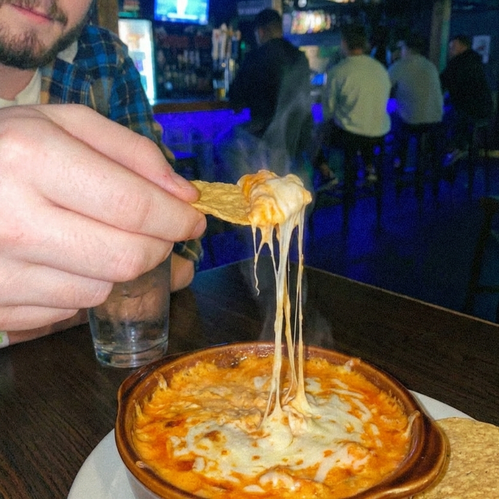 A hand dipping a tortilla chip into a warm buffalo dip with stretchy melted cheese