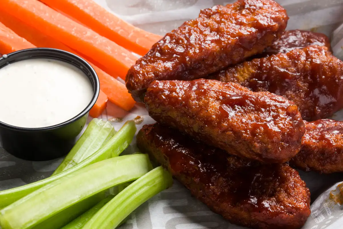 A hand dips a saucy boneless wing into a cup of ranch dressing on a kitchen table