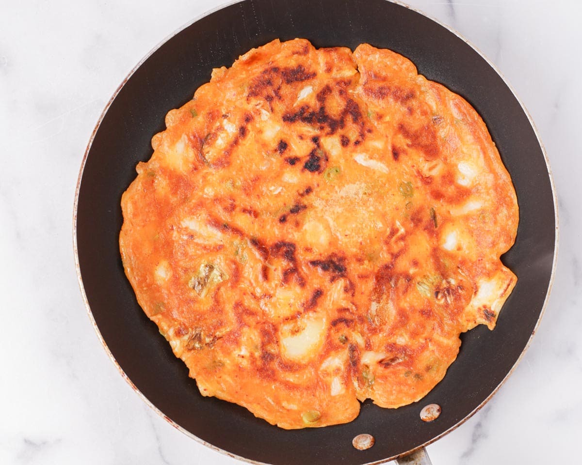 A hand flipping a kimchi pancake in a skillet with a wide spatula, showing a deeply browned underside