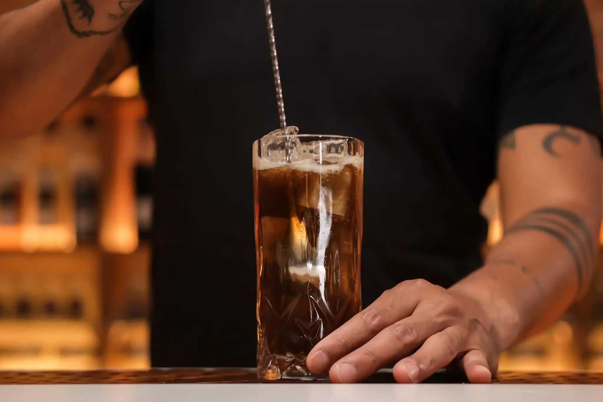 A hand gently stirring a cuba libre with a long spoon in a highball glass filled with ice on a kitchen counter