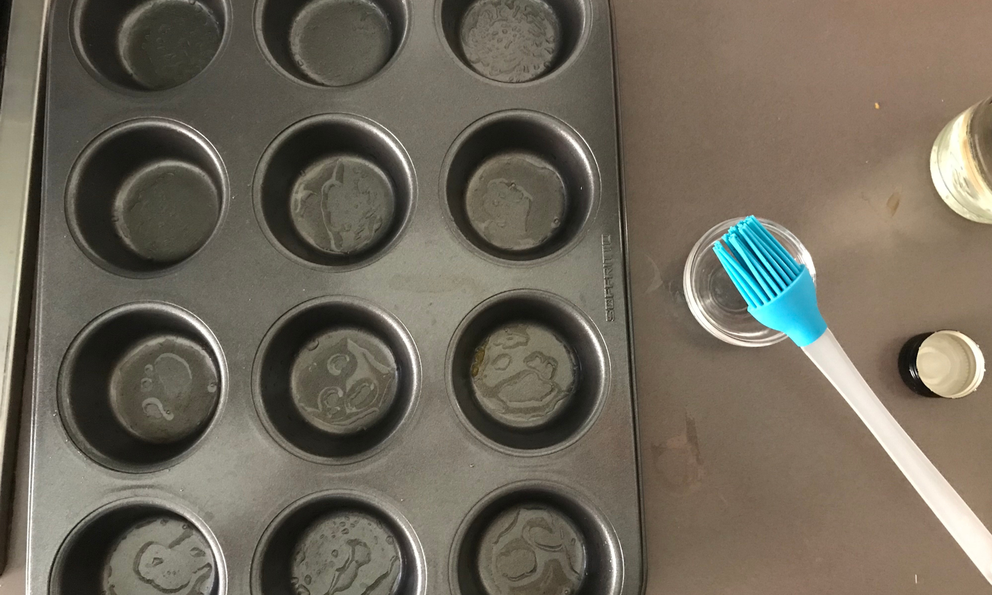 A hand greasing a metal muffin tin with butter using a pastry brush on a kitchen counter