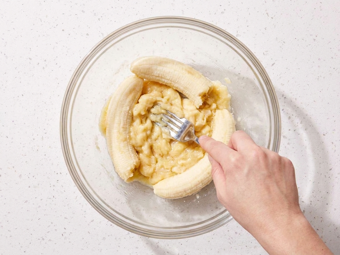 A hand mashing ripe bananas with a fork in a large mixing bowl on a kitchen counter