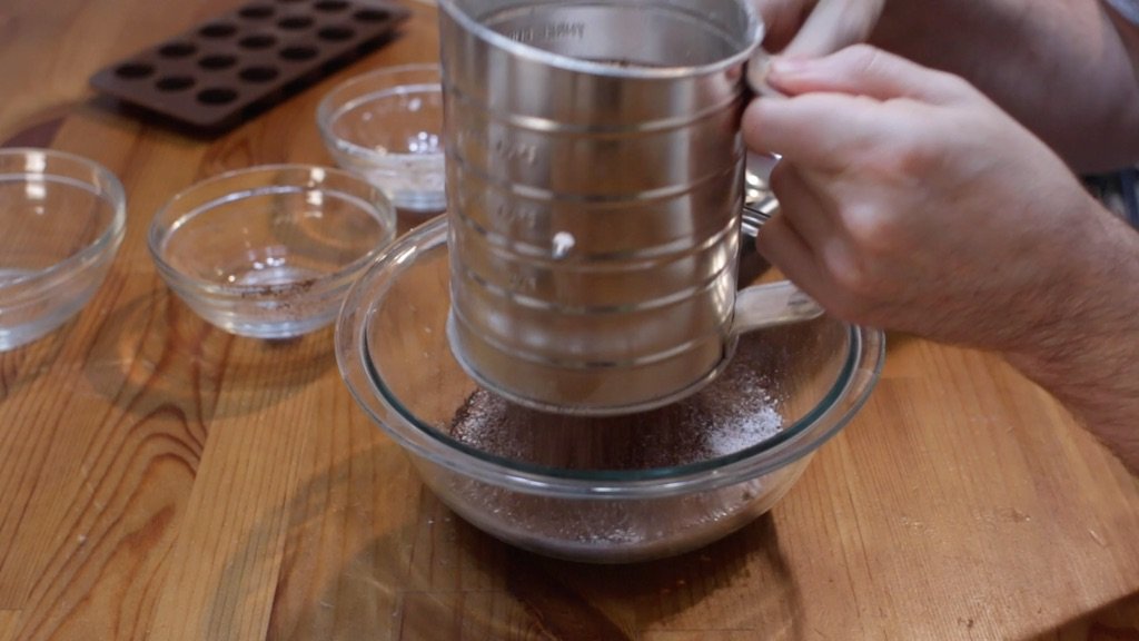 A hand measuring unsweetened cocoa powder into a bowl beside a bowl of granulated sugar on a bright kitchen counter