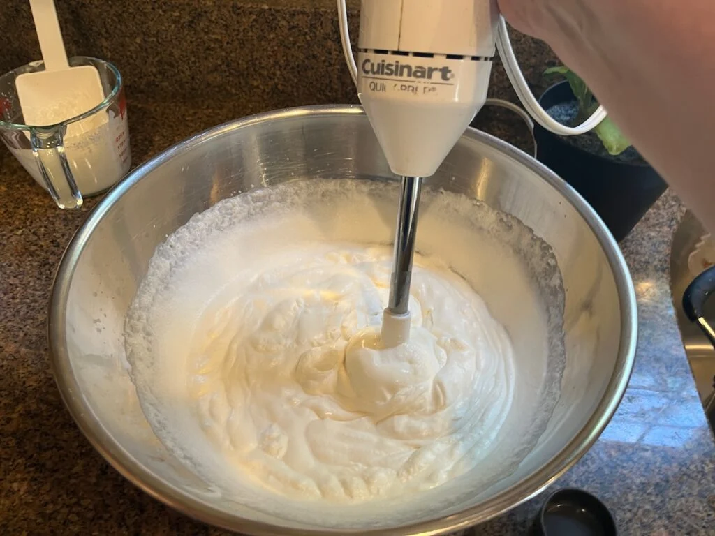 A hand mixer whipping heavy cream in a stainless steel bowl on a kitchen counter