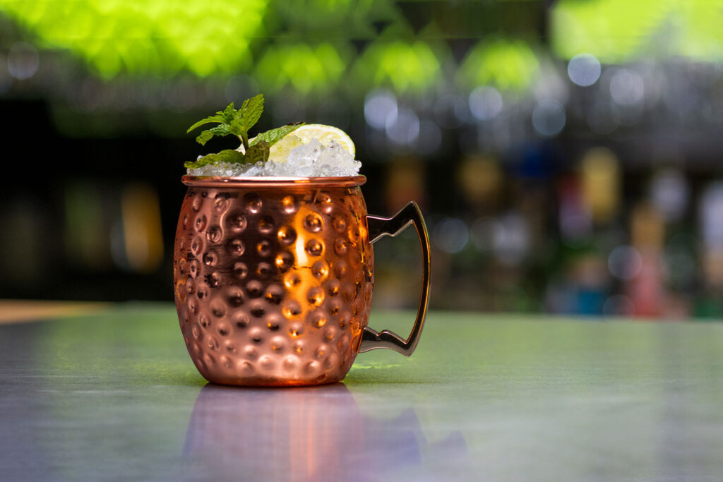 A hand pouring ginger beer from a bottle into a copper mug packed with crushed ice on a kitchen counter