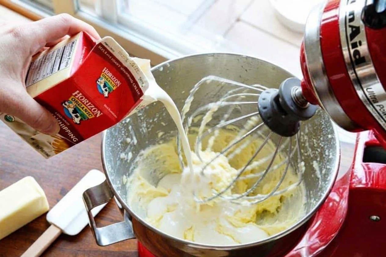 A hand pouring heavy cream into a bowl of buttercream while a mixer runs on low speed on a kitchen counter