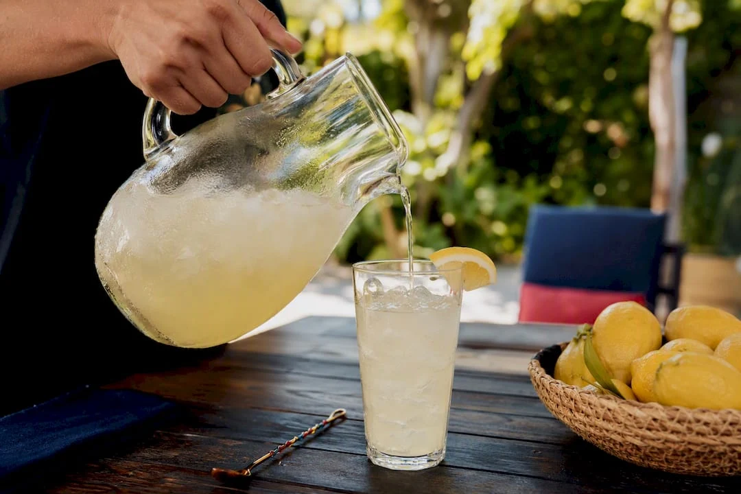 A hand pouring homemade lemonade from a glass pitcher into a tumbler filled with ice on a kitchen counter