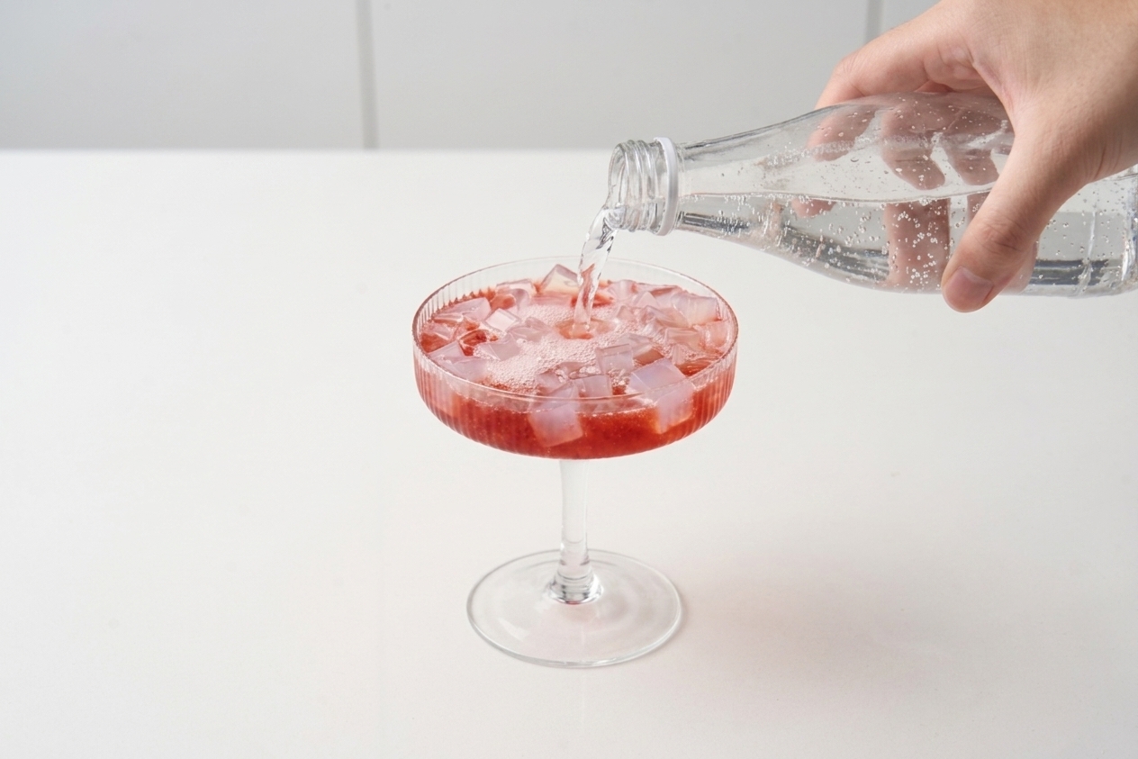 A hand pouring sparkling water into a glass filled with fruit syrup, ice, and konjac jelly pieces on a bright kitchen counter
