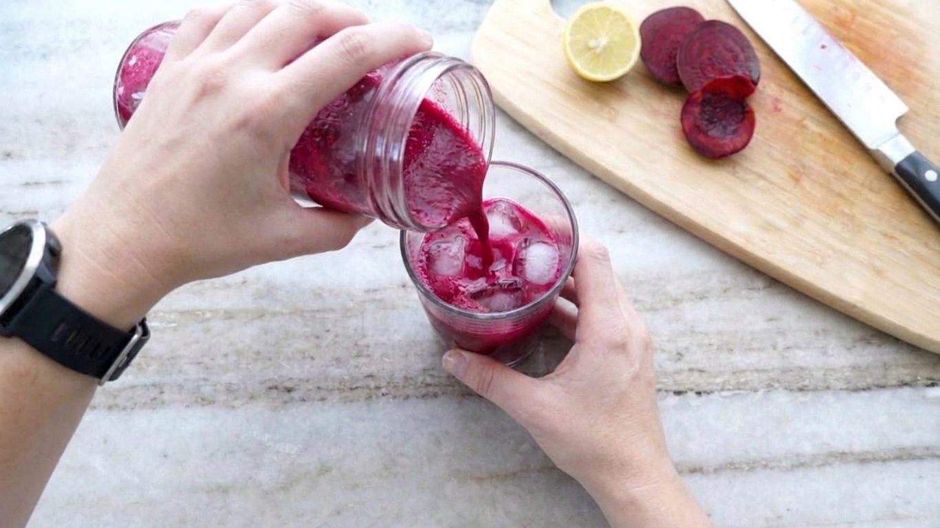 A hand pouring vivid magenta beetroot juice from a glass jar into a tumbler filled with ice