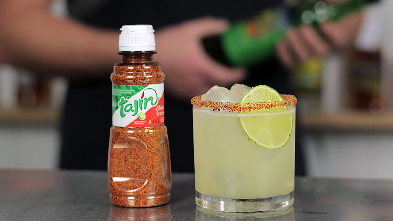 A hand rimming a rocks glass in Tajín and salt on a small plate beside lime wedges on a kitchen counter