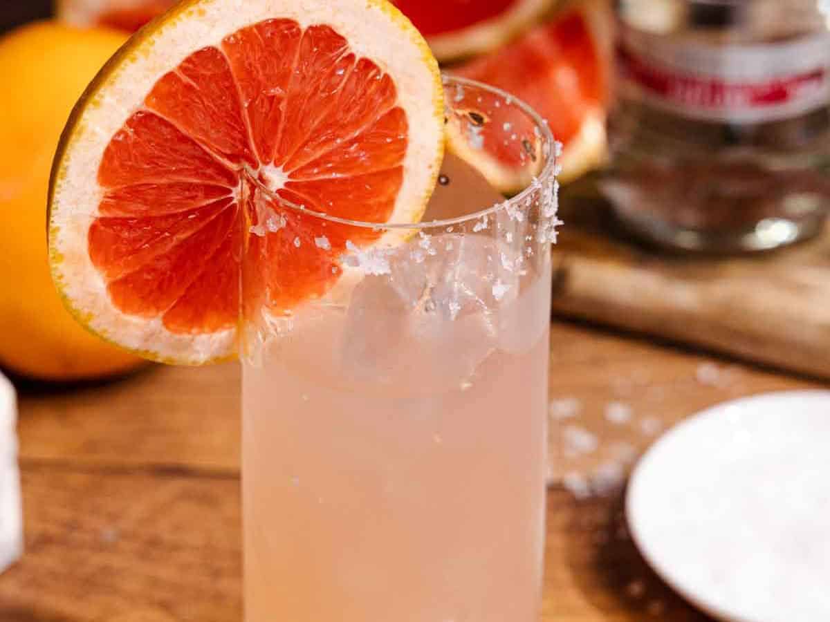 A hand rubbing a lime wedge around the rim of a highball glass next to a small plate of coarse salt on a kitchen counter