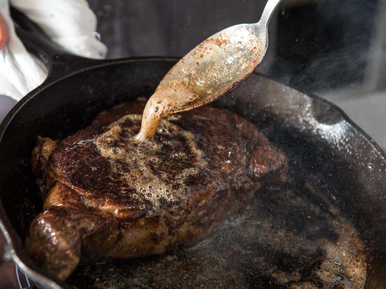 A hand spooning foaming garlic herb butter over a seared filet mignon in a cast iron skillet on a stovetop