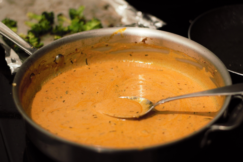 A hand stirring a creamy tomato sauce in a skillet on the stove, with steam rising and a wooden spoon visible