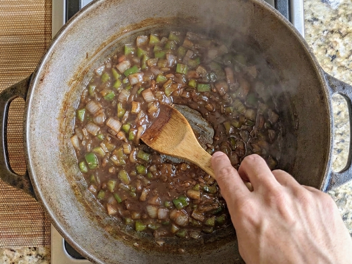A hand stirring a dark roux and vegetables in a heavy Dutch oven on a stovetop, with a wooden spoon and steam rising